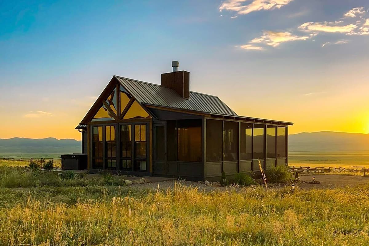 A cabin near Westcliffe, Colorado, stands in an expansive valley meadow with the shining sun and towering peaks in the backdrop.