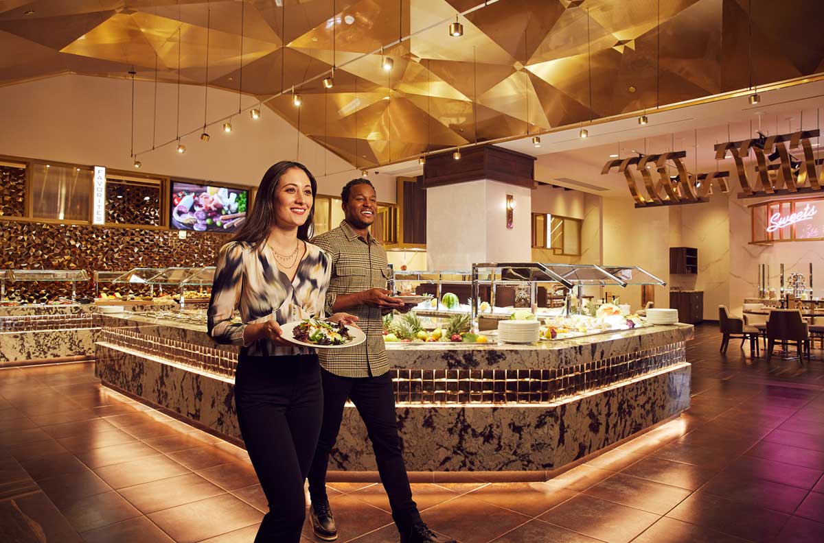 A couple carry loaded plates from a stocked buffet. They are very pleased with themselves