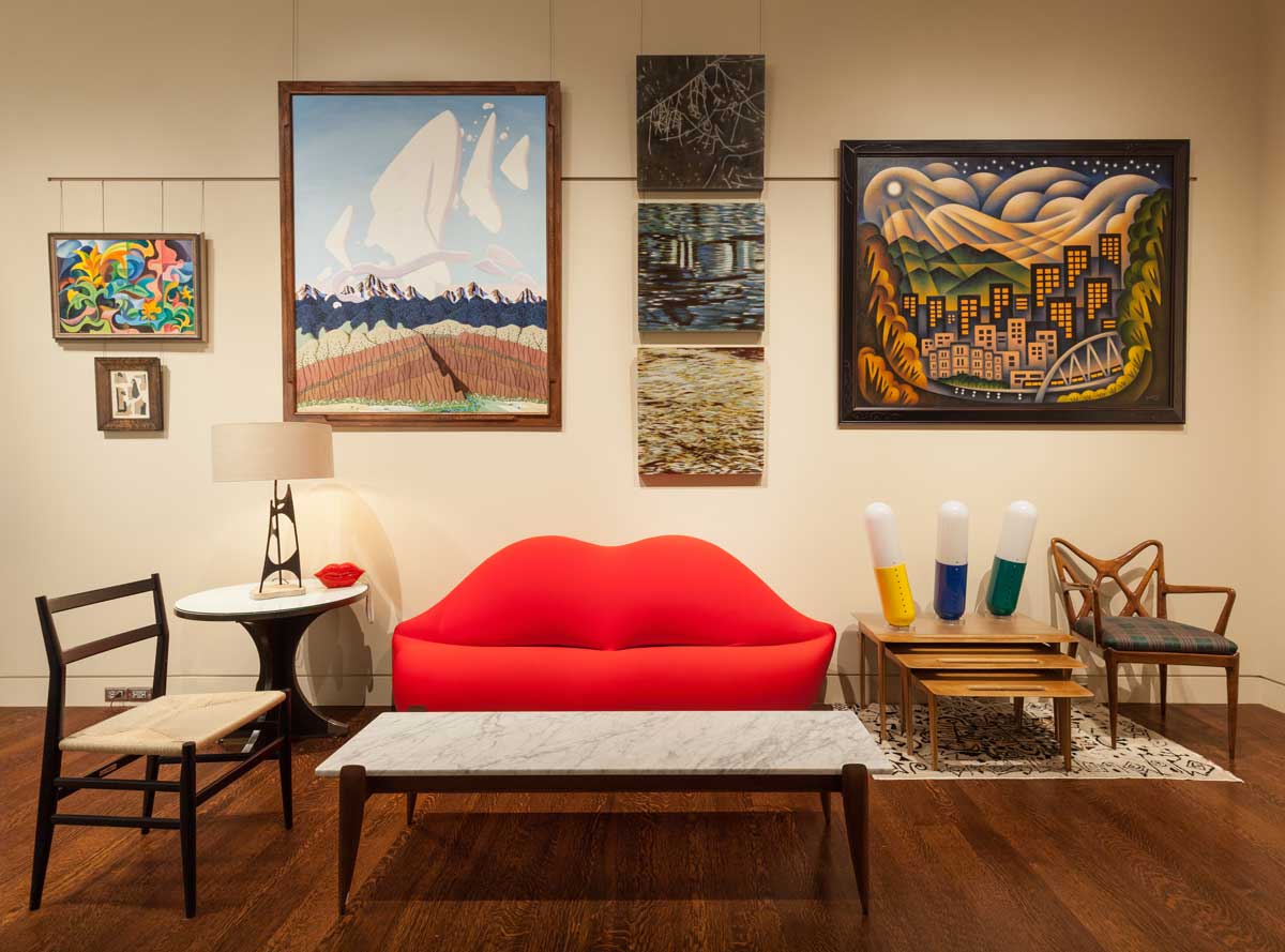 A living room exhibit at The Kirkland Museum of Fine & Decorative Art, which includes eclectic paintings and photos on the wall, mismatched furniture and a bright-red couch in the shape of lips.