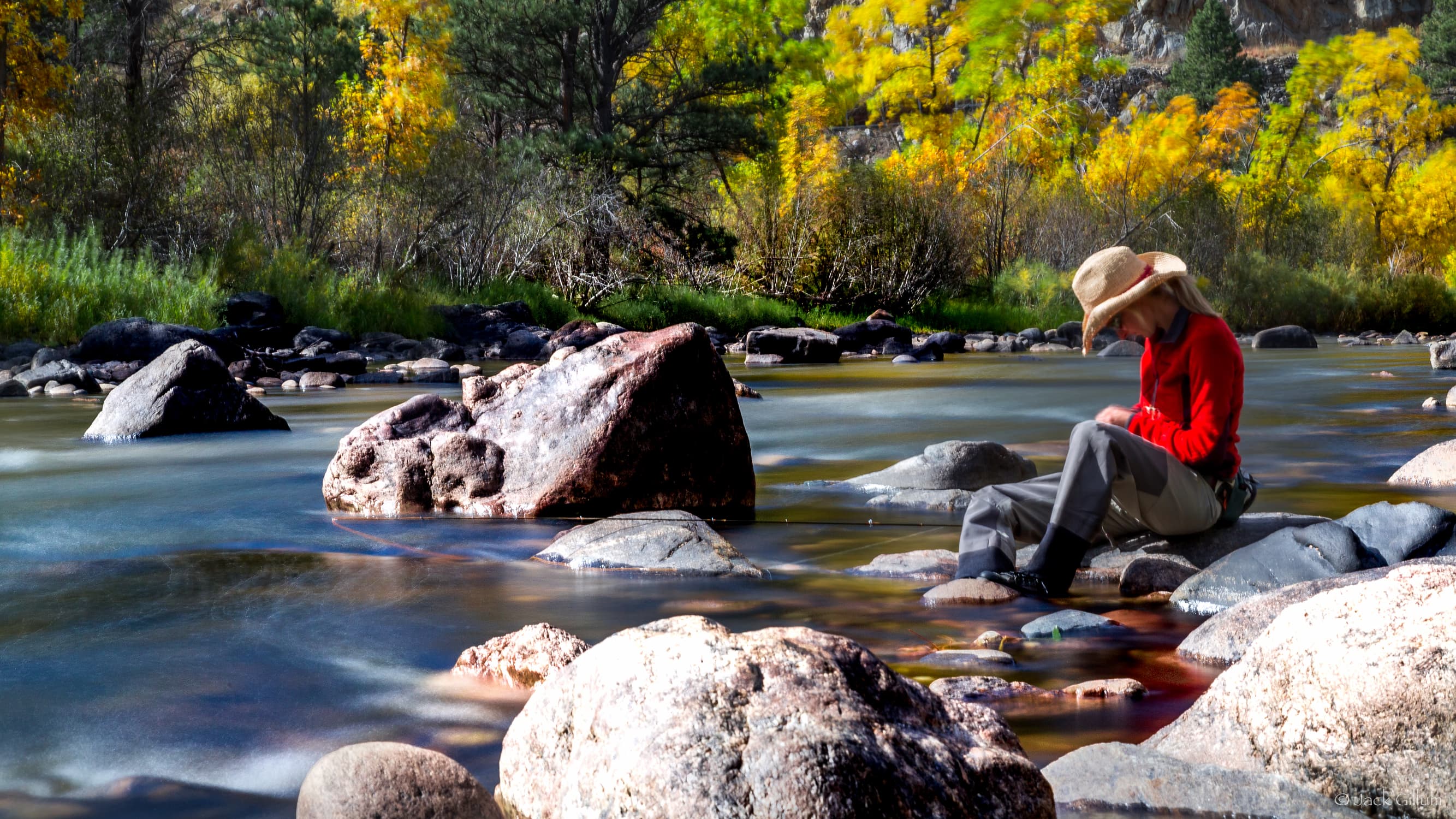 A fisher wears waders, a red sweater and a wide-brim hat and sits on a rock near the shore of the Cache la Poudre River.