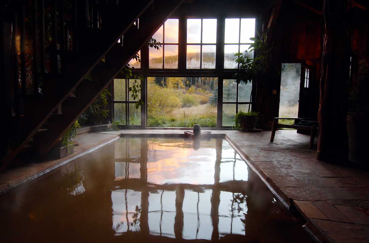 A person soaks in the steaming waters of a bath house at Dunton Hot Springs. They rest their arms and head on the stone floor and look out the wall of windows.