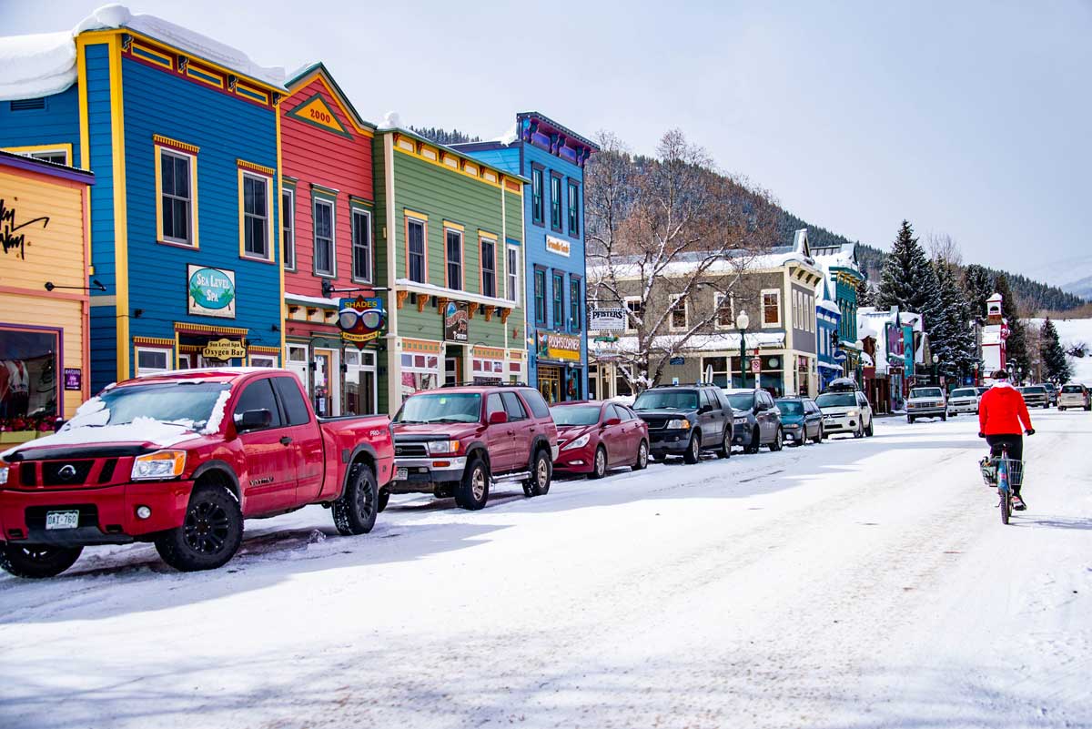 Blue, pink, green and yellow storefronts line a snoy street. A cyclist pedals up the street and cars are parked along the side