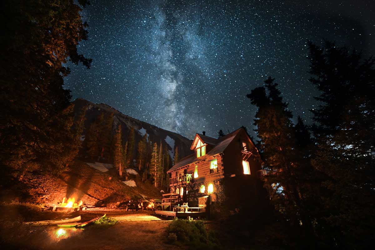 A three-story log cabin at Alta Lakes, Colorado, stands next to a crackling fire surrounded by a pine forest. Above is a sky full of sparkling stars.