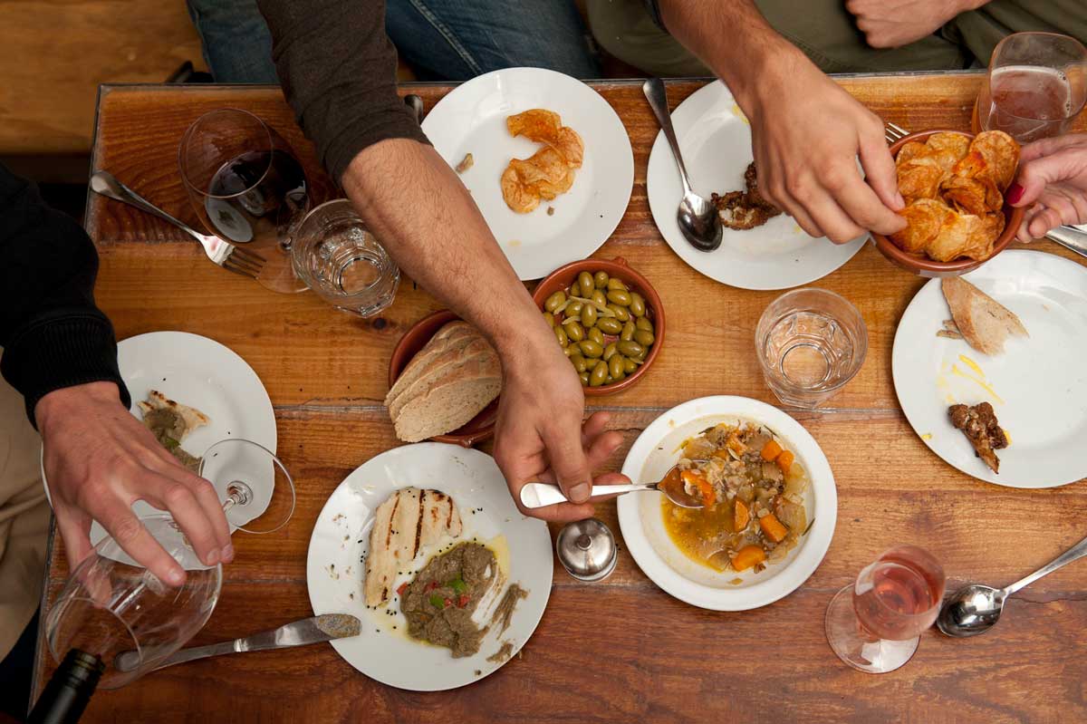 Four diners' hands reach for small plates of cooked meats and veggies at Cafe Aion in Boulder in a birds-eye view of the wood table.