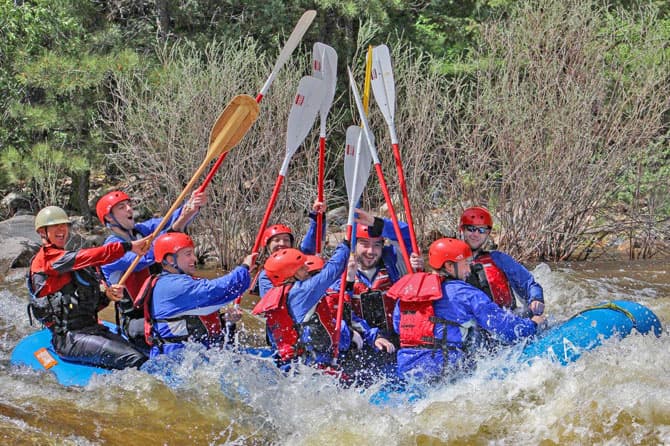 A large group of red vest- and helmet-clad people are on an inflatable blue raft on the white waters of the Cache la Poudre River in Colorado. They are all holding their paddles up in the air in celebration as they cruise down the river. Behind them are various green plants.