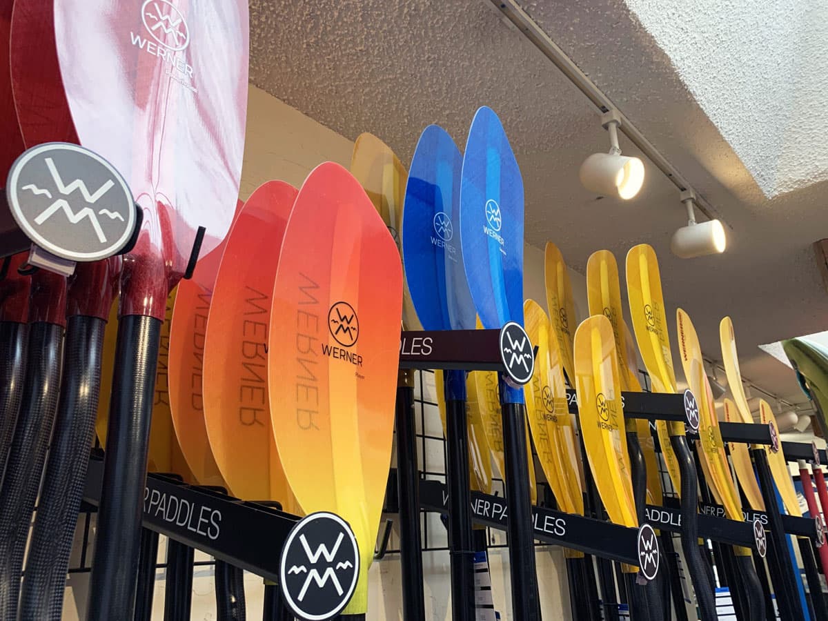 A close-up image of red, orange, yellow and blue water sports paddles hung upright on hooks against a beige wall. The paddles are labeled, "WERNER" and are staggered along the wall. Above them are popcorn ceilings and two small spotlights.