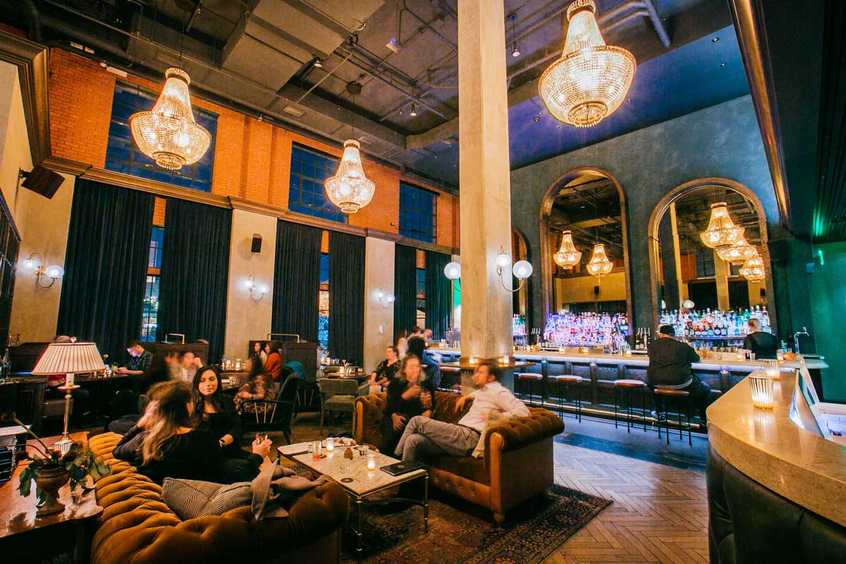 Bar patrons in a cozy, jewell-toned bar relax on plus couches with glowing chandeliers above them