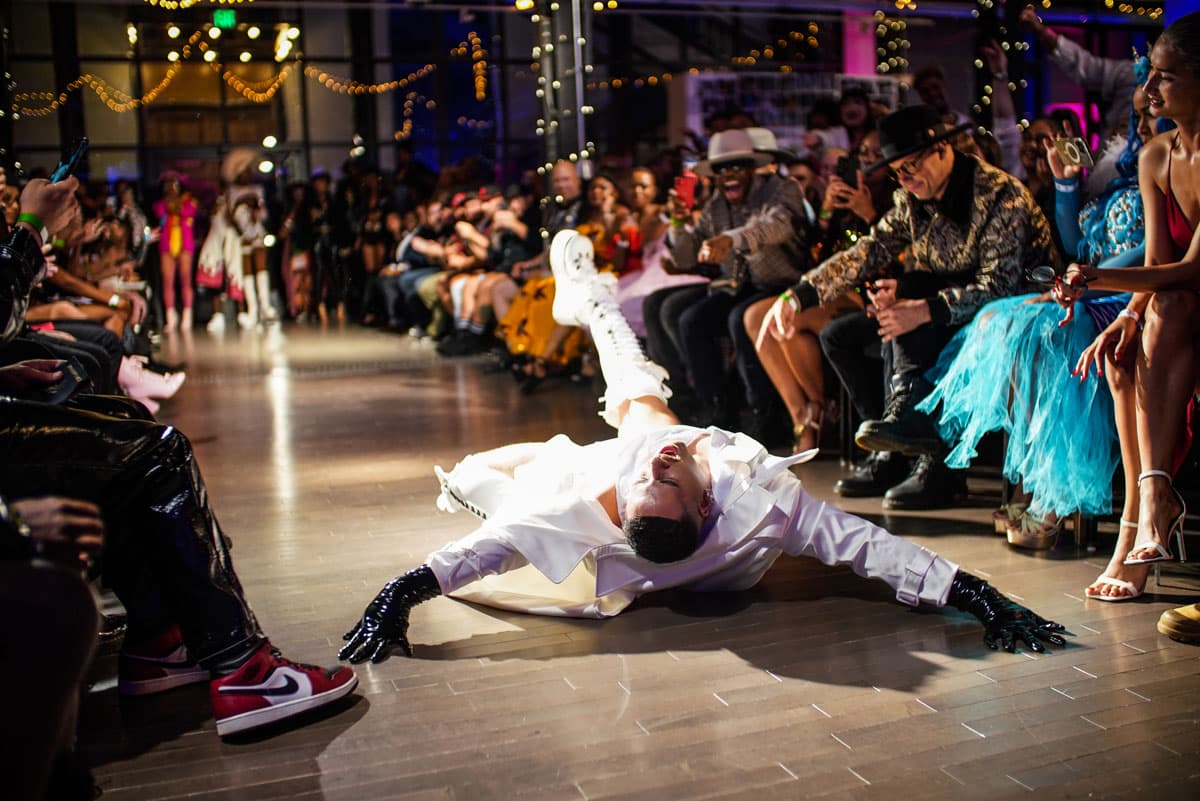 A person in a white trench coat, thigh-high laced white platform boots and black gloves is mid-death drop in the center of a fashion walkway in Colorado.