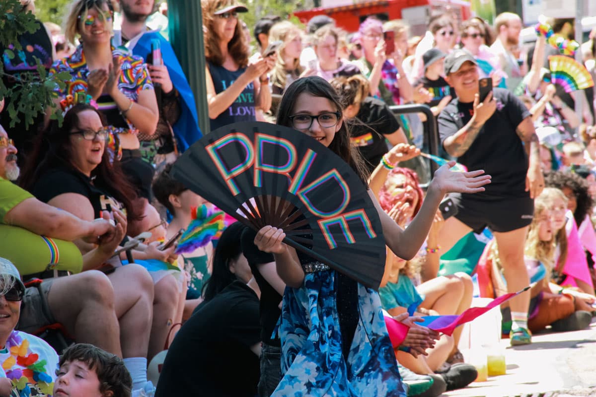 A smiling child holds a black folding fan that says "PRIDE" in all caps, rainbow letters. Around them a crowd of smiling and laughing parents and children watch a parade.
