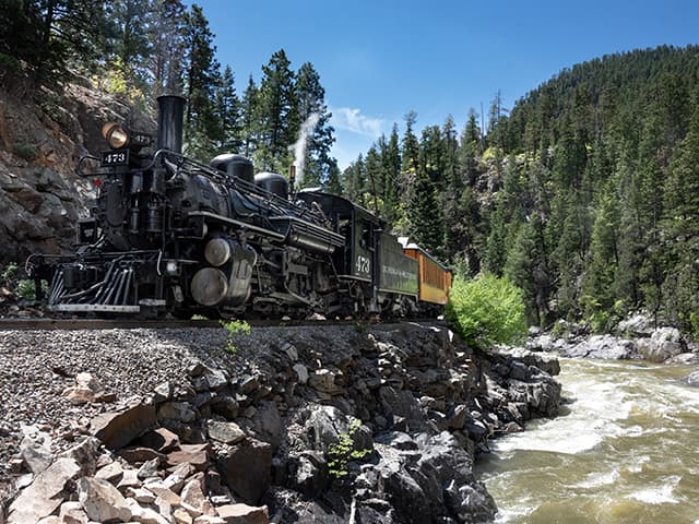 The Durango Train chugs along its track beside a rushing river. Along the sides of the tracks are small cliffs of sharp rocks. In the background is a large hill with pines.