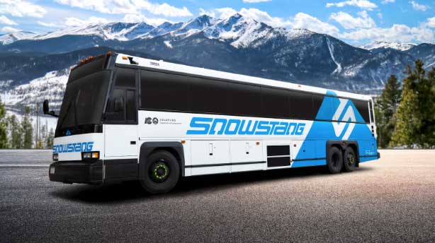 A public transit bus with Snowstang on the side parks, ready to go. Snowy mountain peaks rise behind it
