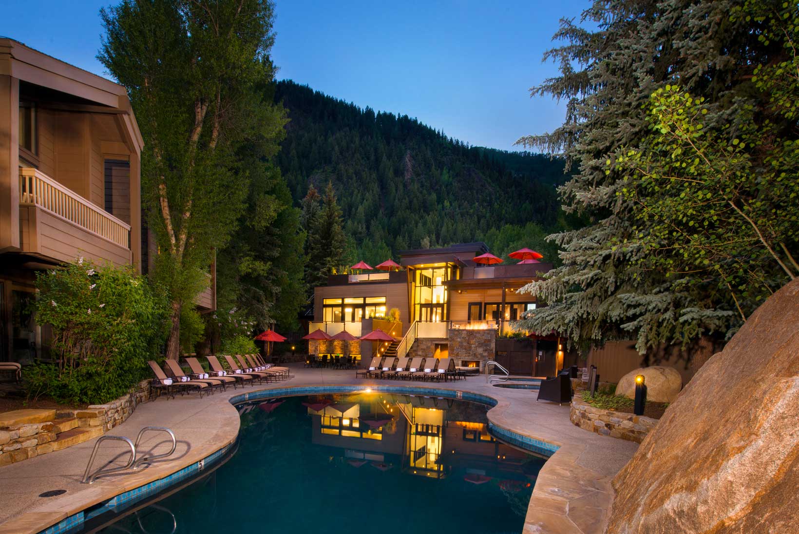 Night falls over a hotel pool, with the lit-up lodge and evergreen forest in the distance