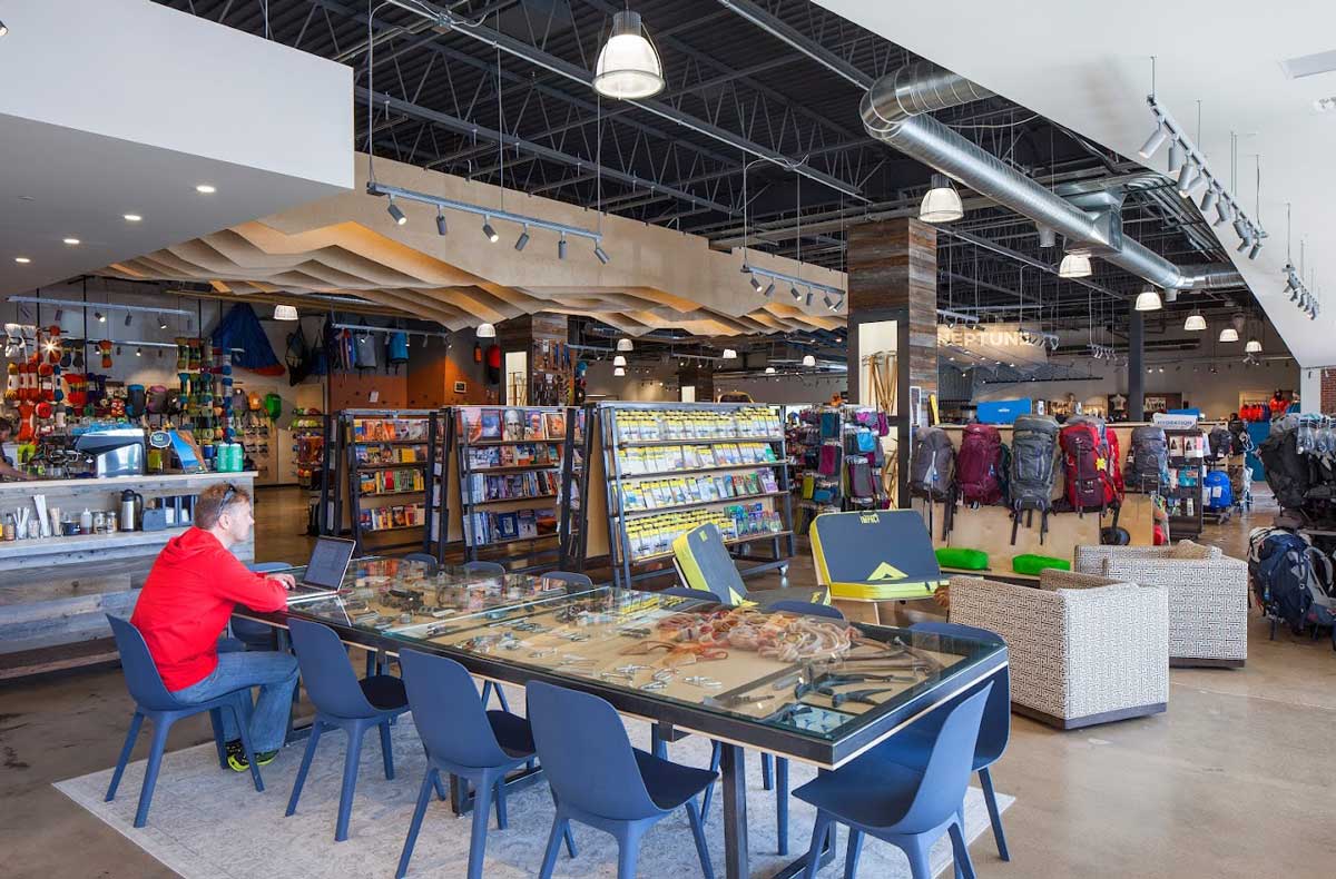 A person works from a laptop in store packed with outdoor gear, including backpacks, maps, and climbing equipment