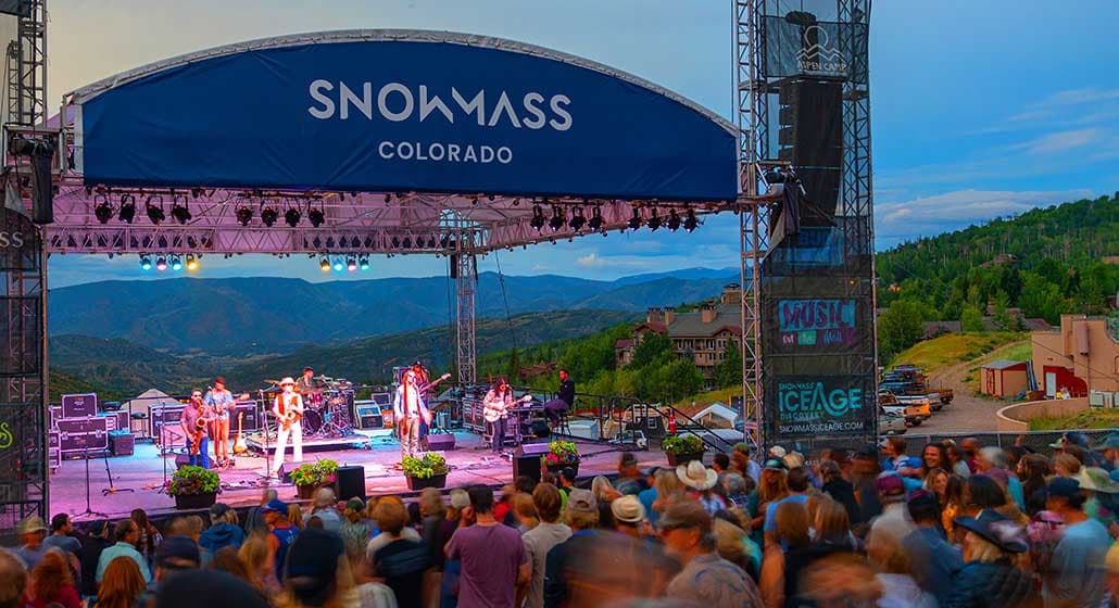 A crowd gathers on the mountainside in front of a stage where a band plays as the sun sets