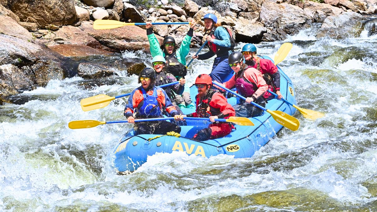 A group of seven rafters hold their paddles and are dressed in protective gear on a blue inflatable raft with the Arkansas Valley Adventures Rafting & Zipline logo. The rafters hold up their paddles victoriously and smile as the raft rolls down white-capped waves.