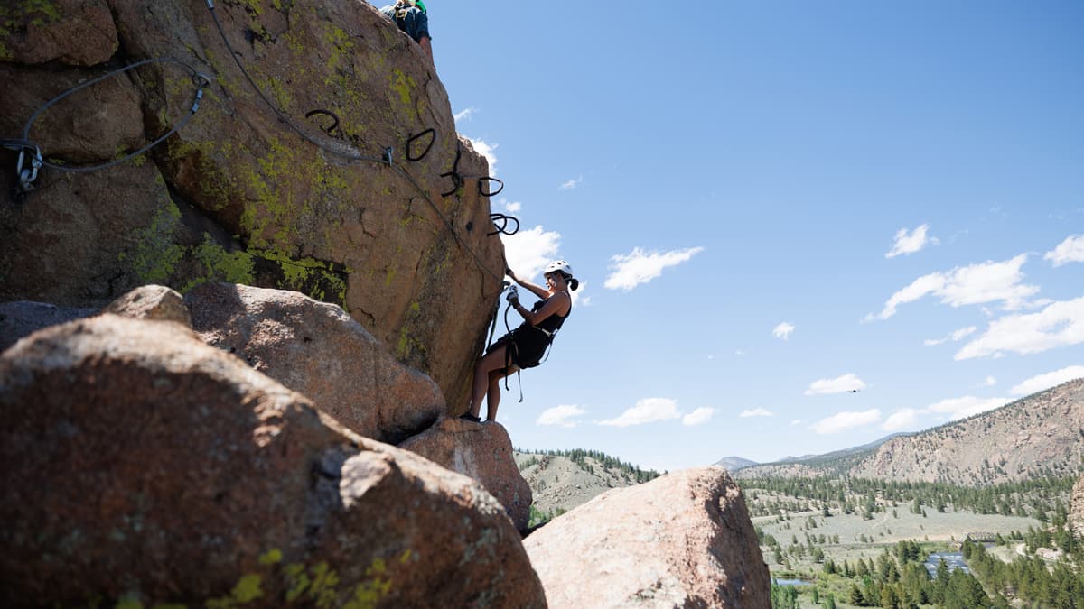 A climber in a white helmet tethered safely traverses the rock climbing route with Arkansas Valley Adventures Rafting & Zipline. The sky above is almost cloudless and dusty blue, and the land sweeping around the climber is vast and mountainous, dotted with trees.