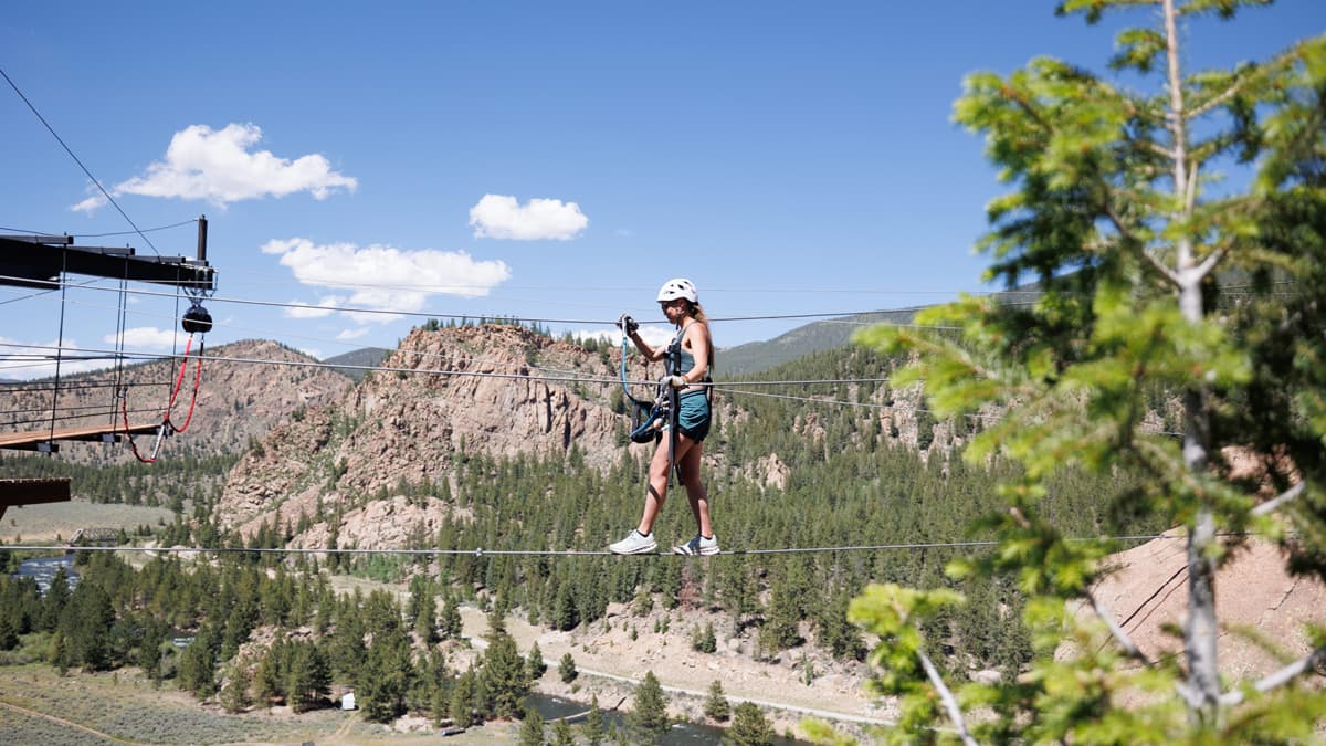 A person wearing a helmet walks across a line from one platform to another in Buena Vista, part of AVA Rafting & Zipline's adventurous offerings. The sky above is nearly cloudless and the craggy mountains in the distance are peppered with tall deep-green trees.