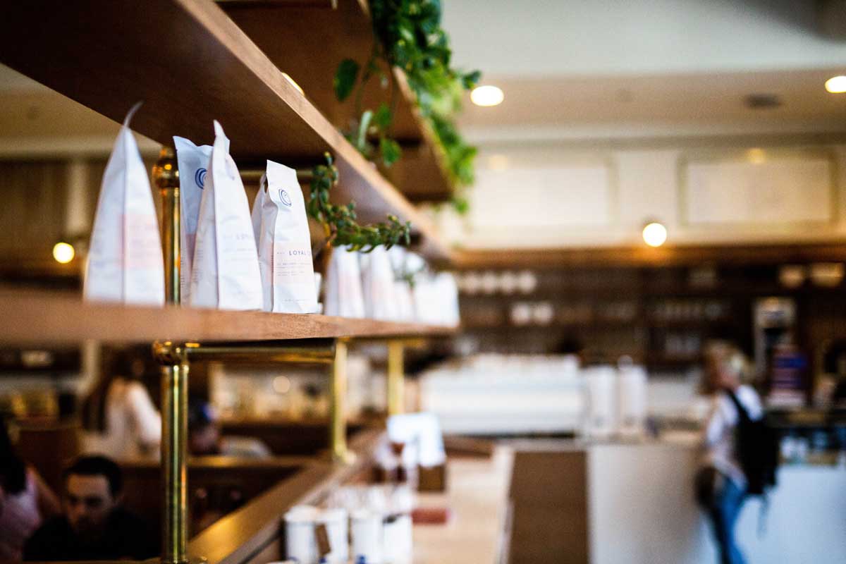 A shelf of bags of ground coffee lead to a barista's coffee bar