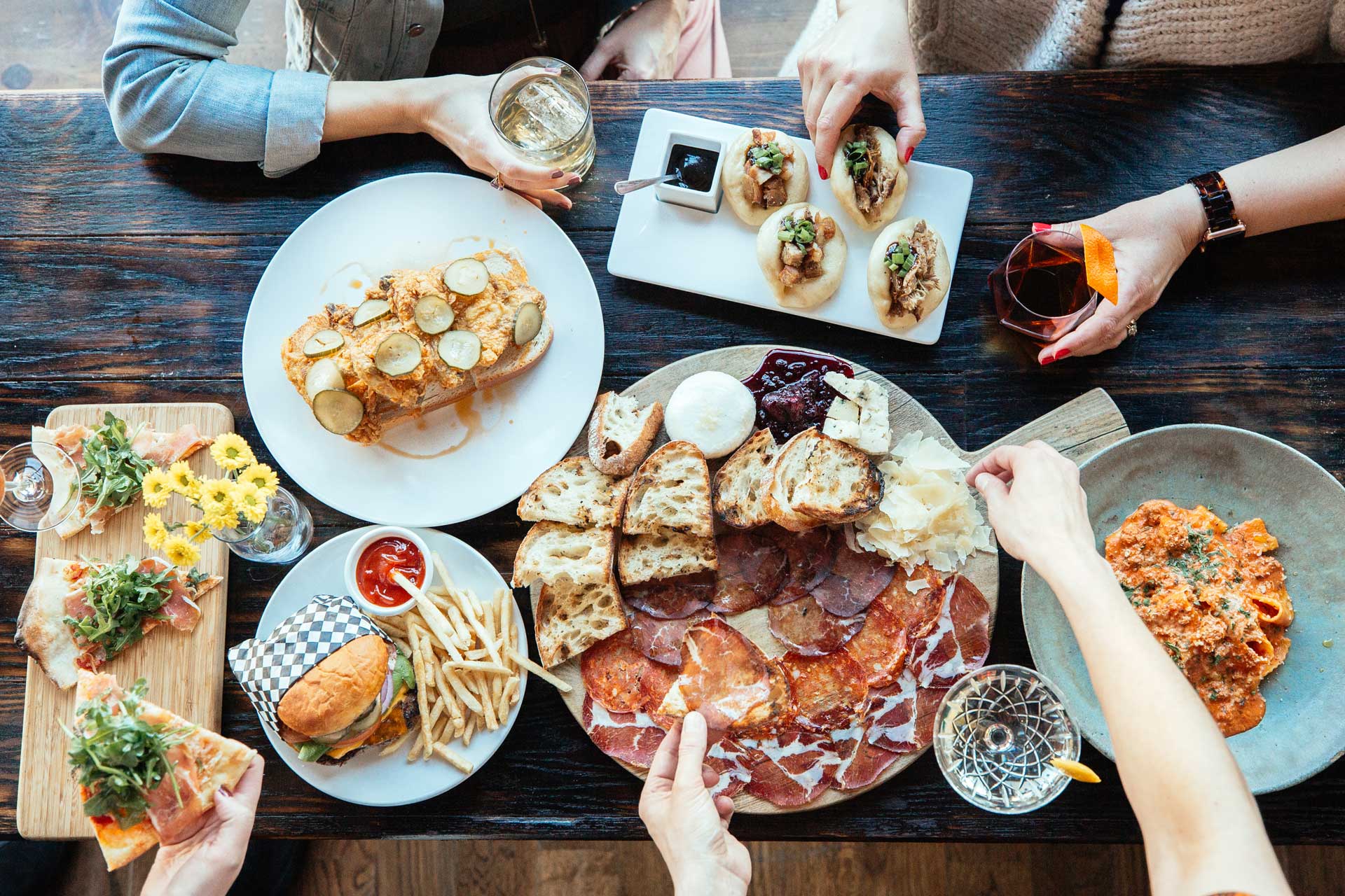 A birds-eye view of a table filled with dishes and hands grabbing burgers, tacos, meats and cheeses off of them
