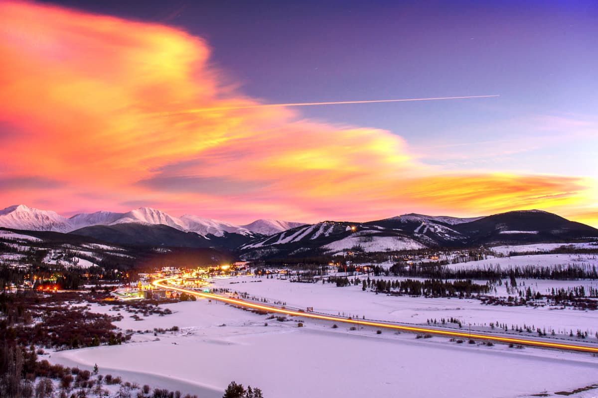 A panoramic image of the mountains of Winter Park, Colorado, under a setting-sun sky, paintedin neon, wispy orange clouds. There is snow everyone on the ground and the lights from the city glimmer in the night sky.