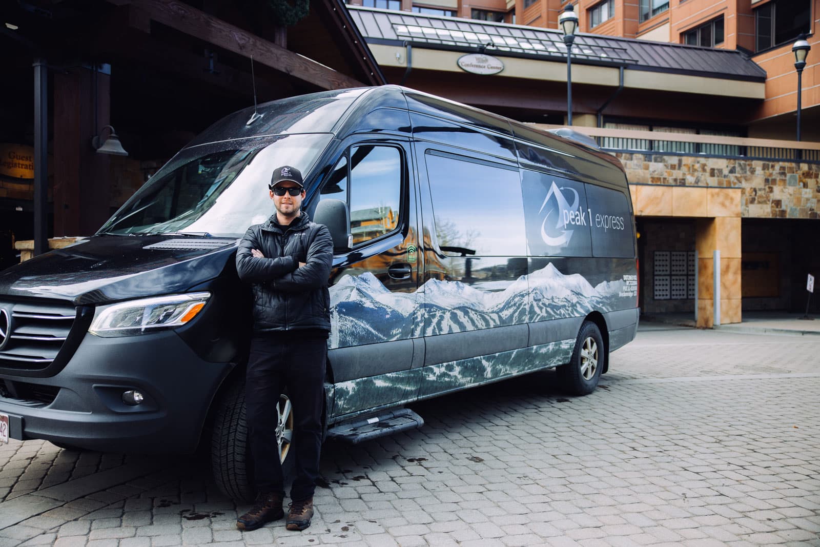 A black van is parked in front of a hotel entrance; the driver stands in front of it smiling