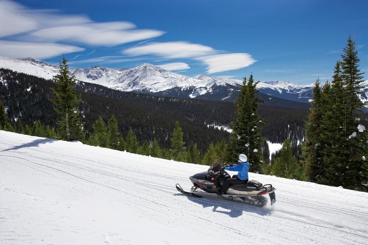 Someone dressed in warm attire zooms along snow-blanketed terrain on a large snowmobile. To their right is a drop off of the hill, which opens up to a pine-tree-laden view underneath snowcapped peaks. The sky above is mostly clear and deep blue.