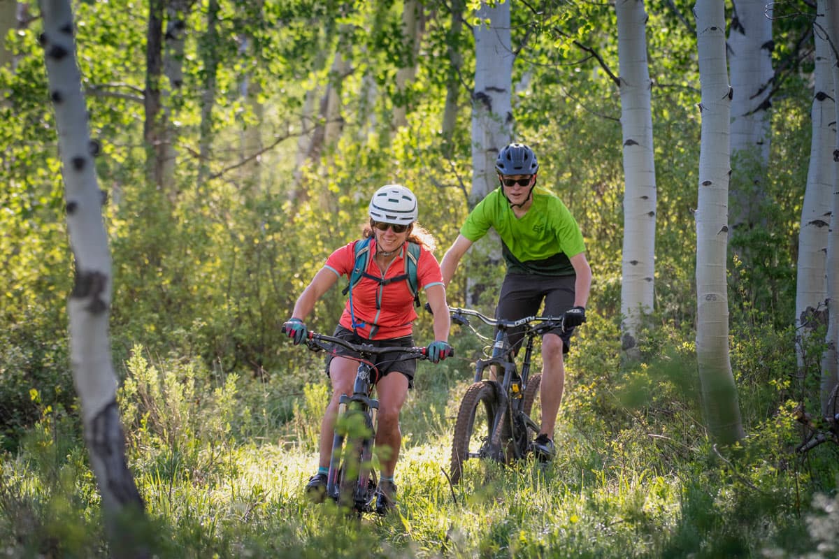 Two smiling people wearing sunglasses, helmets, and outdoor athleisure gear bike through the aspen trees in a grassy uphill field.