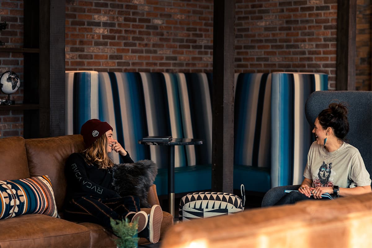 Two people sit in the communal space at The Bivvi Telluride in Colorado a laugh together. The room features brick walls, a striped corner nook and thick leather couches with accent pillows.