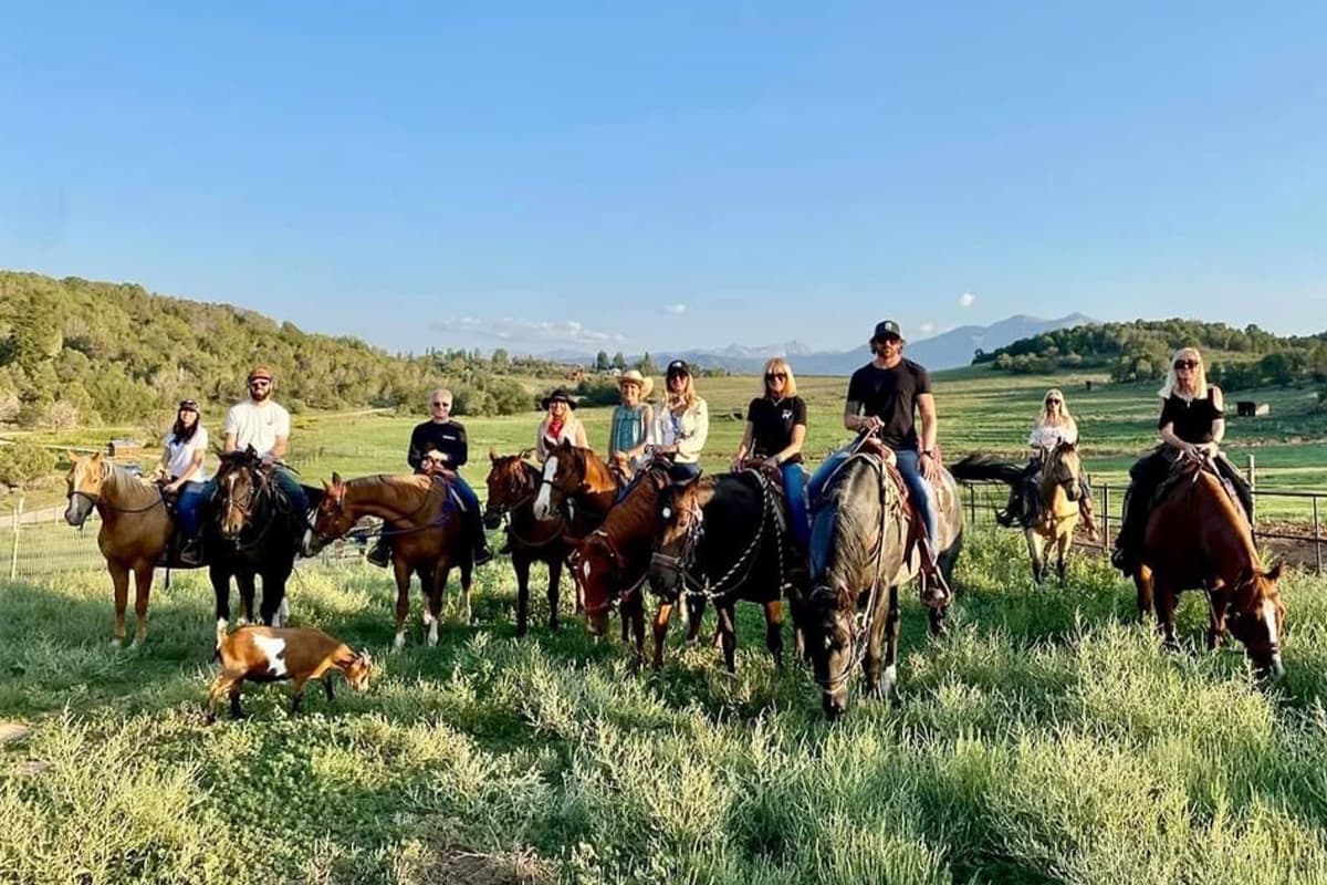 A group of riders at Cedar Ridge Ranch sit atop horses in a rich, green field and pose for the camera. A curious goat wanders into the photo.