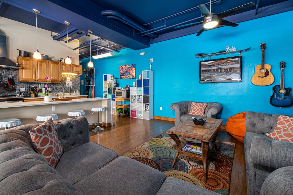 A large room with one wall painted bright blue and the ceiling painted dark blue. To the left of the photo is a kitchen with cabinets, barstools, an oven, counter and sink. On the wall next to the kitchen is a bookshelf and a microwave, and next to that is the living area with three cray couches with accent pillows. On the bright blue wall straight ahead are several paintings and two acoustic guitars, one of them a deep blue color.