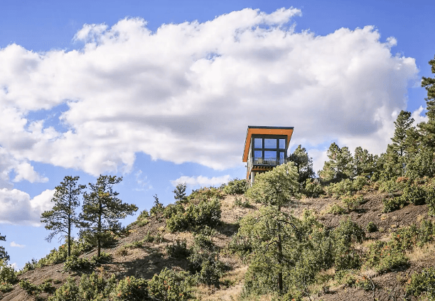 The Ridge cabin in Pagosa Springs perches on a shrub-covered ridge with blue sky and white, fluffy clouds in the background.