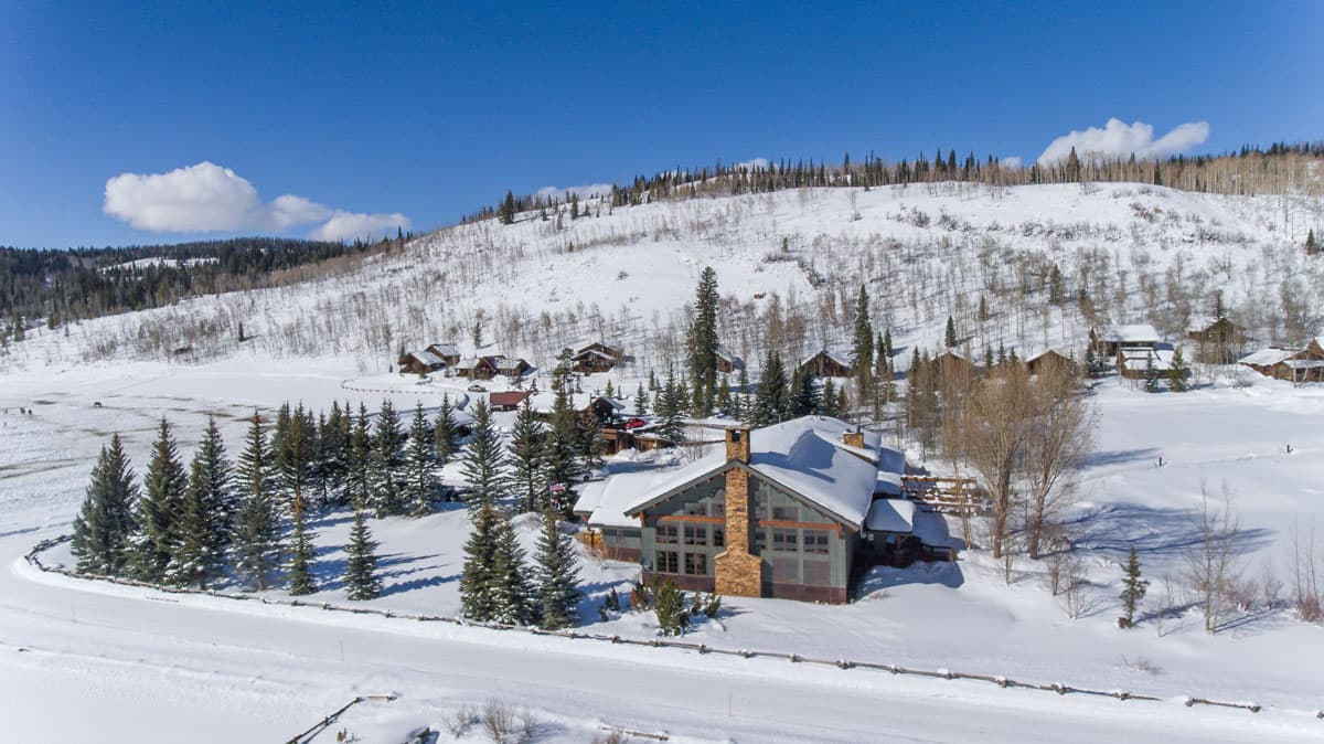A deep blue sky sits above a large snowy and tree-covered hill in Clark, Colorado. At the base of the hill is a large clump of wood cabins with snow on their roofs, surrounded by trees. At the forefront of the photo is a larger cabin with a broad, tall stone fireplace.