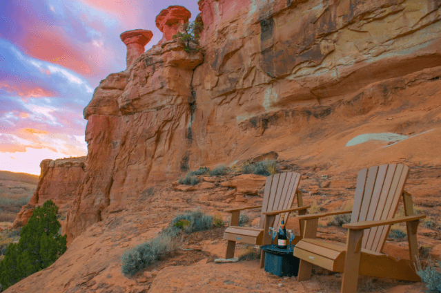 Two Adirondack chairs sit under red-rock grandeur and a colorful sunset
