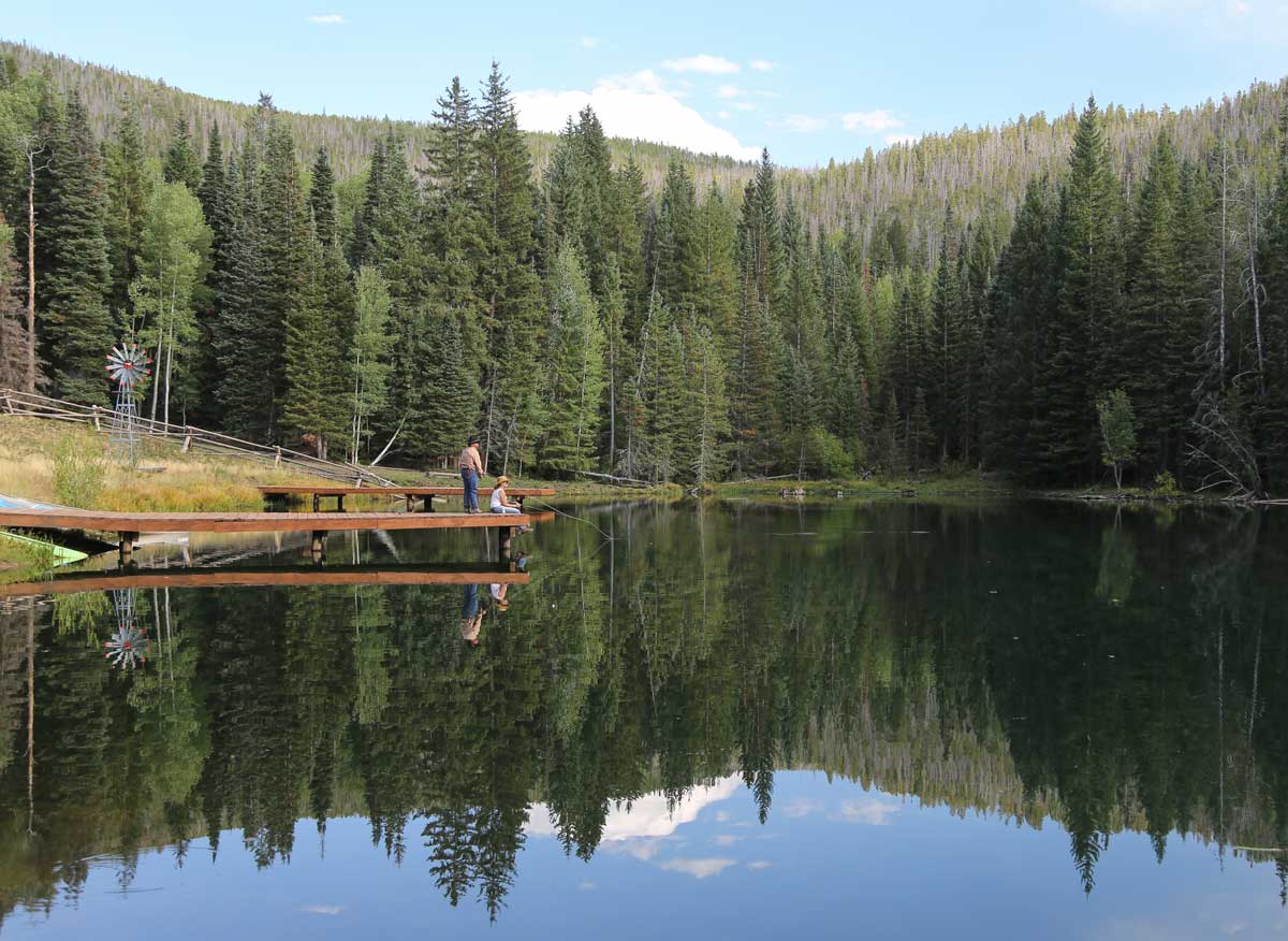 Anglers fish from a pier as mountains and evergreen trees reflect in the water