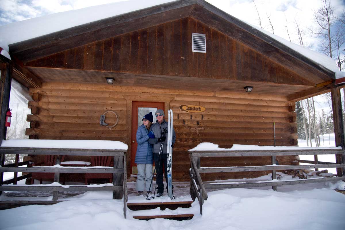 Two people wearing warm coats stand on the snow-covered front steps of a cabin at Latigo Ranch holding Nordic skis