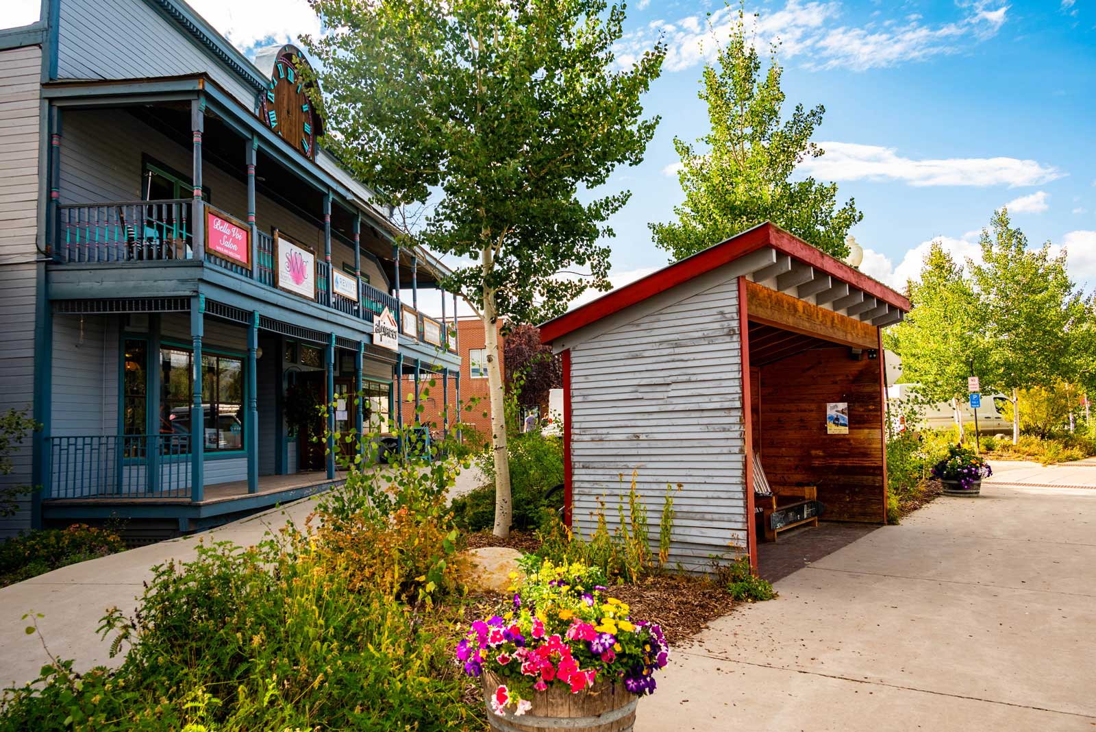A wood-framed bus stops sits in front of a few houses with balconies; the sun and flowers are out