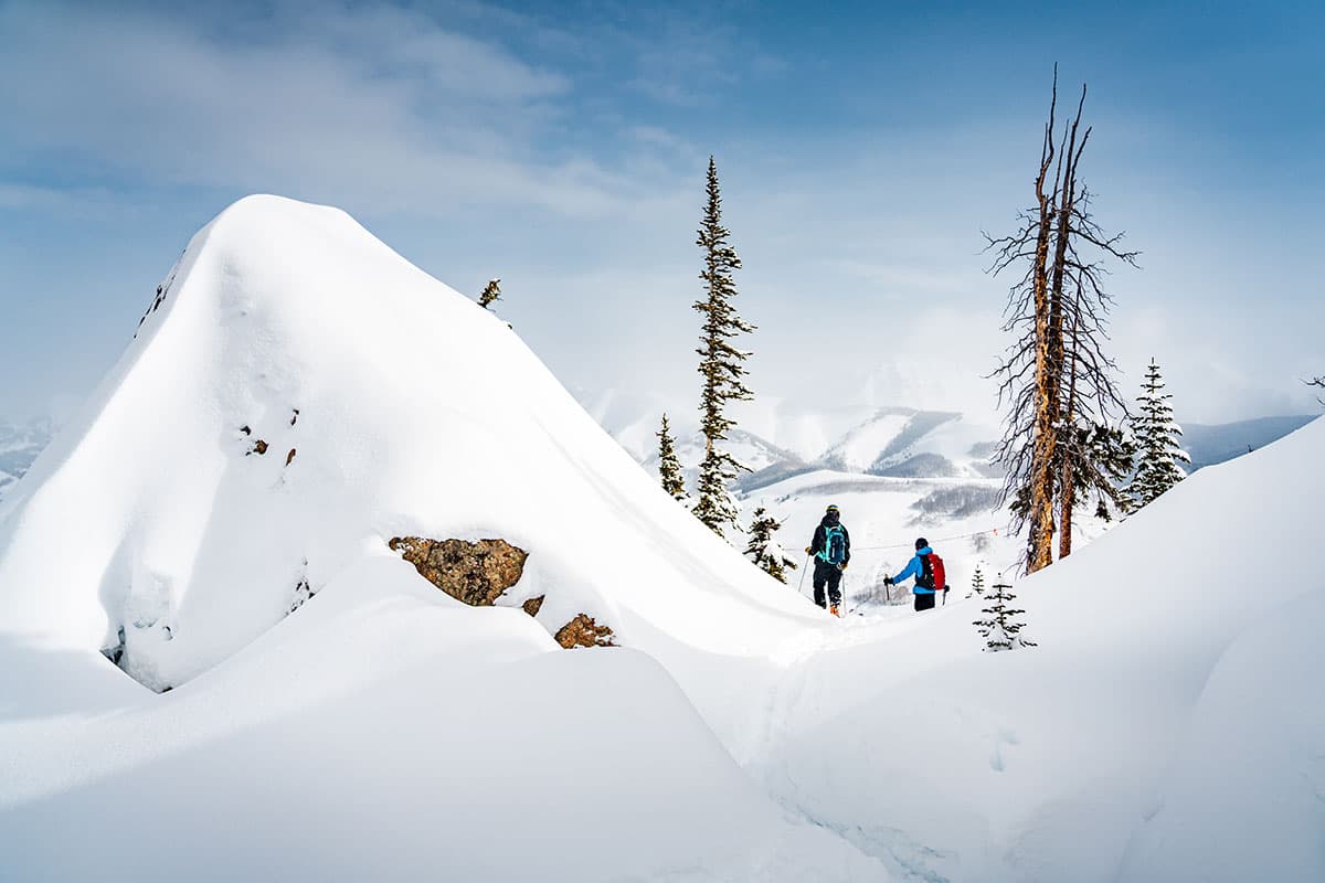 Snow-covered rock with two skiers behind it and mountains in the distance