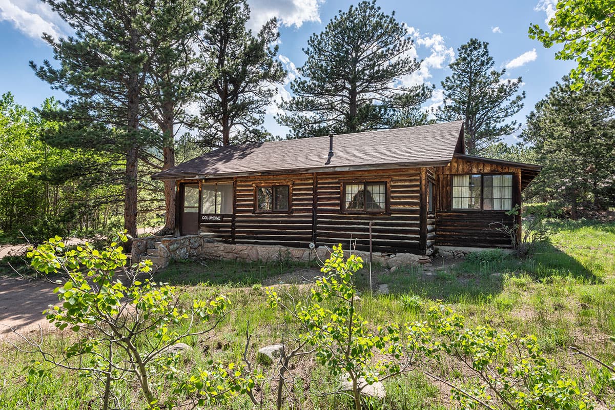 A log cabin at Meeker Park Lodge sits on a lovely wooded lot with tall pine trees and a cloud-streaked blue sky behind it.