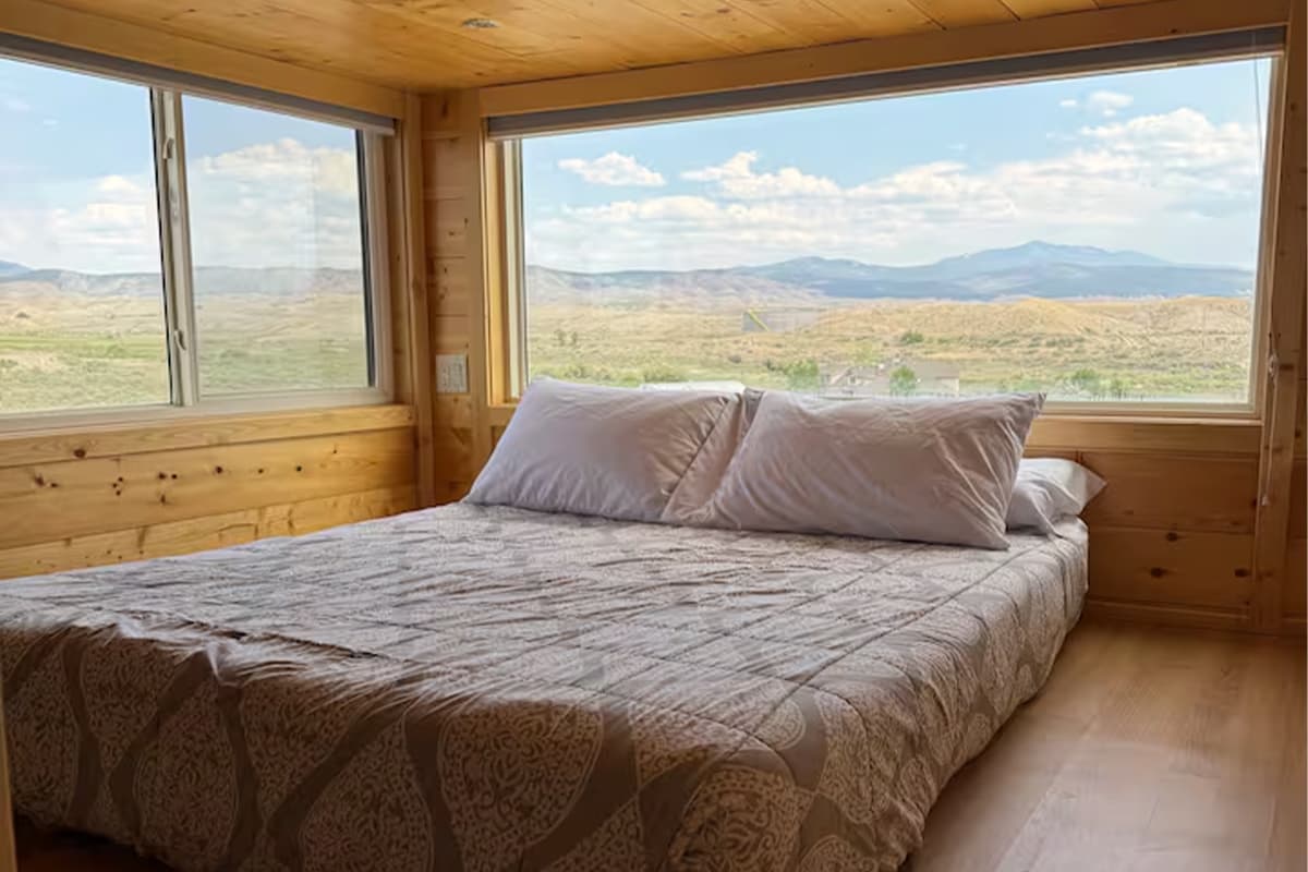 In a light-wood-lined bedroom, a simple bed rests in front of several windows offering panoramic views of Montrose, Colorado.