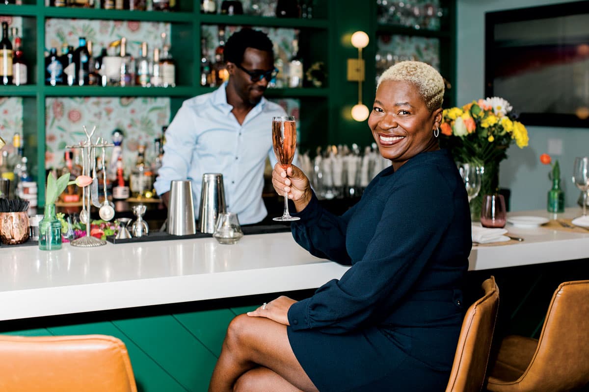 Mawa McQueen sits at the bar in her Aspen restaurant cheersing the camera with a glass of champagne