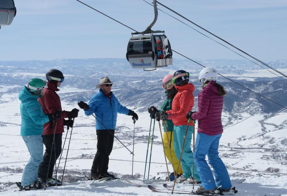 Five skiers in colorful gear listen to another skier at Steamboat. In the background a gondola makes it's way up the mountain on a winter's day. The ground beneath them is covered in snow.