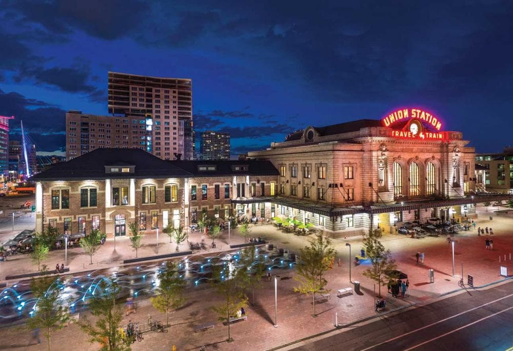 Denver's Union Station at night. The historic building's red neon sign reads "Union Station" above a clock and below the clock it says "Travel By Train." There are small green trees surrounding a square with a water feature. There are people mingling around the station. And the blue night sky features some high rise buildings behind the building.