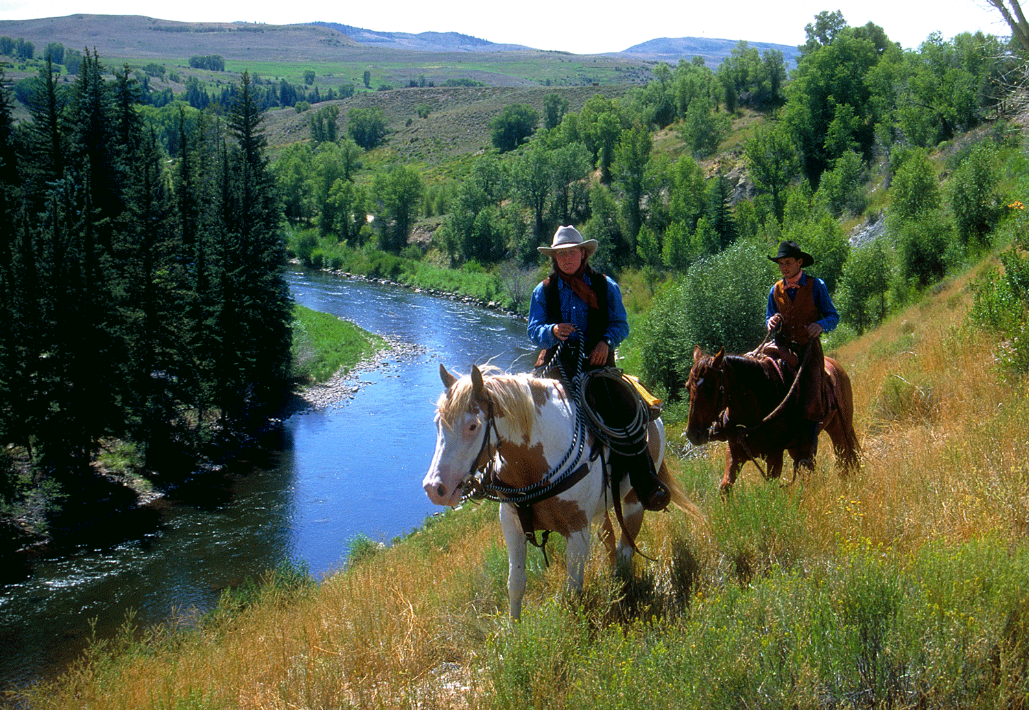 Two people ride horses alongside a still blue river
