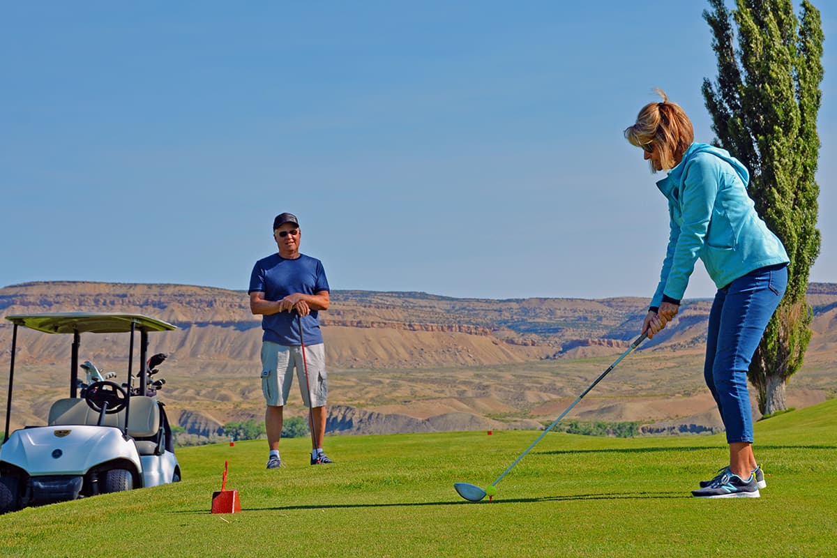 A woman gets ready to swing at a golf ball on a green as a man watches on during a blue-sky day.