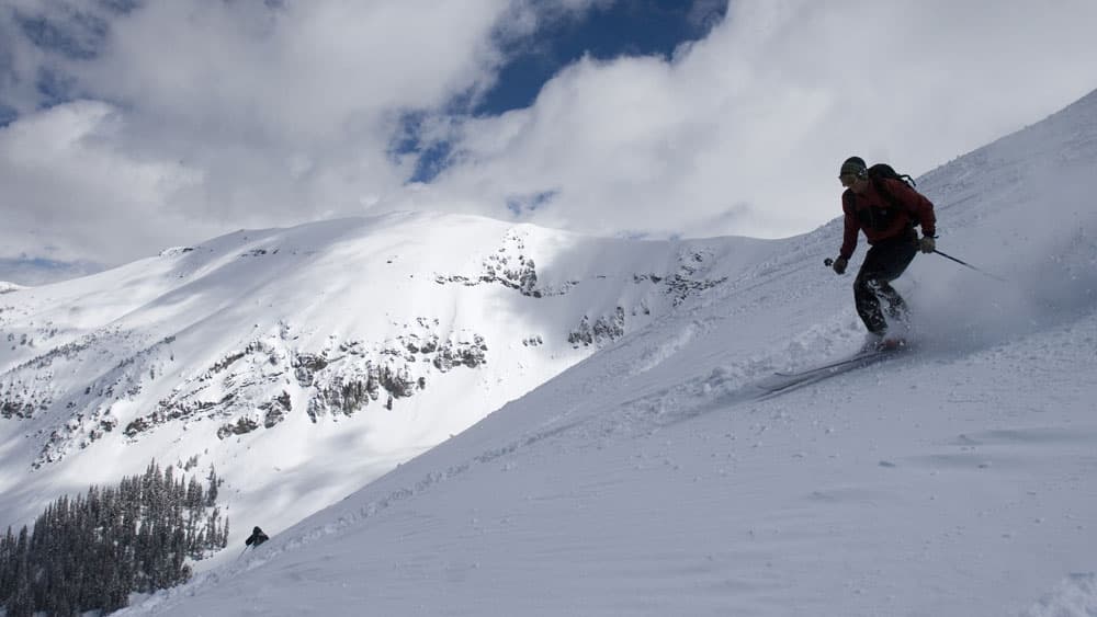 A skier in dark clothing kicks up fresh powder near the top of a snow-covered mountain ridge at Telluride Ski Resort in Colorado.