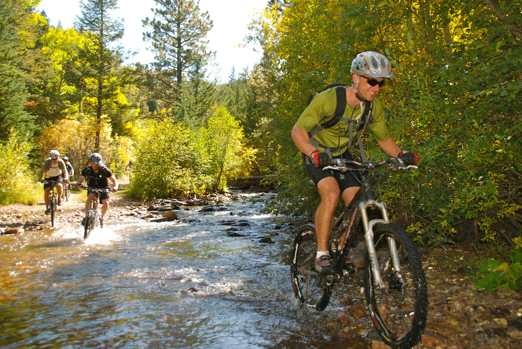 Three smiling and be-helmeted mountain bike riders churn their legs through what appears to be a flowing creek. Lots of green aspen trees line the banks for the creek.