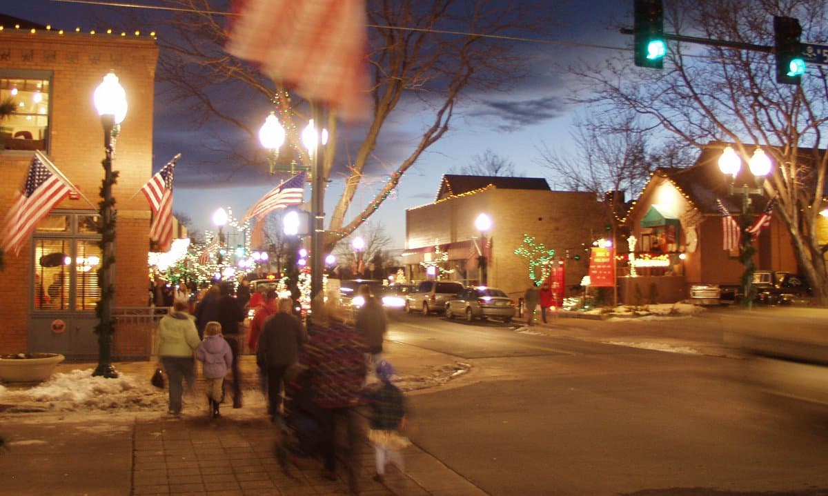 Olde Town Arvada bustling at night with many people walking the streets, shopping, dining and meeting with friends. The ground has snow and there are Christmas lights on buildings.