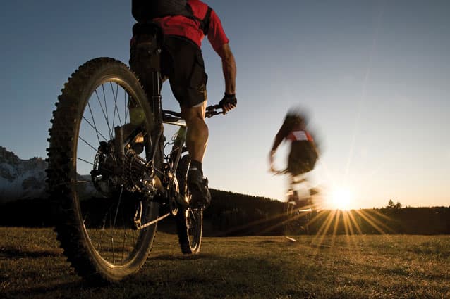 The sun rises up over the distance horizon and illuminates two cyclists mountain biking through a grassy field in Colorado.
