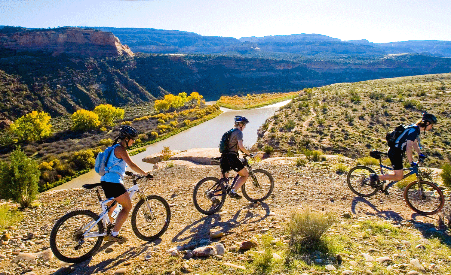Three mountain bikers crest the top of a mesa with a panoramic view over the mighty but brownish Colorado River, with many mesas of different heights rising in the distance