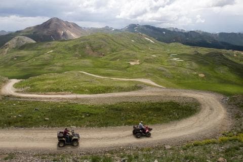 Two ATVers zoom down a winding dirt road split with green-grass fields. In the distance are jagged peaks under a cloudy sky in the San Juan Mountains.